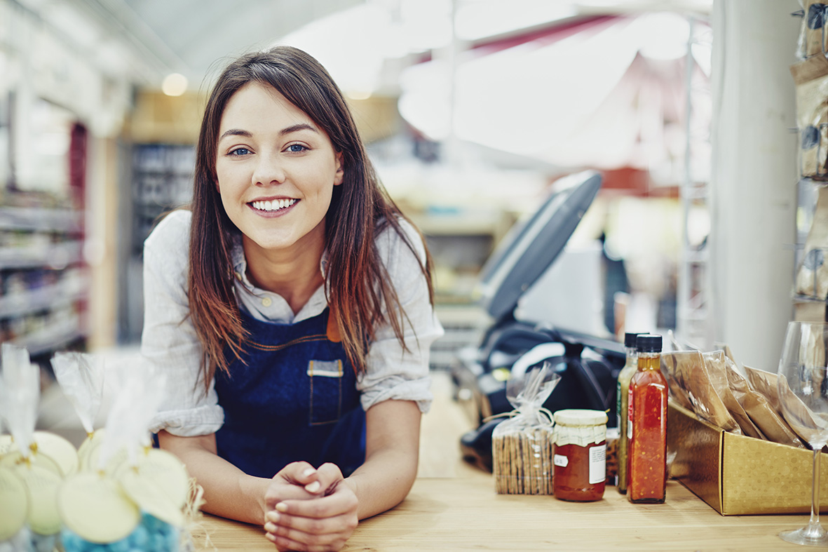 lady smiling behind a counter with a till and some groceries on the table