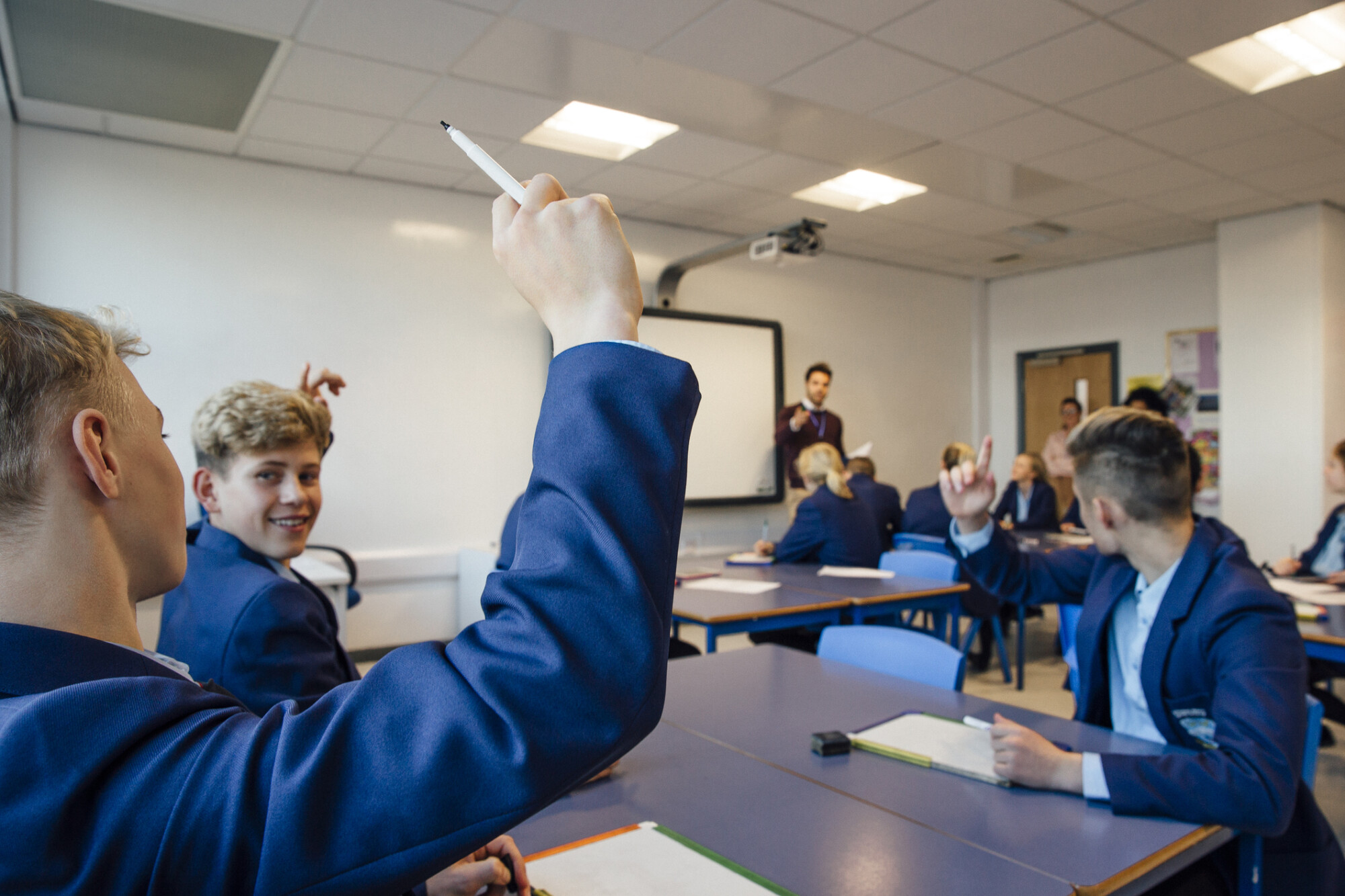 Point of view shot of a high school lesson where the teacher has asked a question and some students have their hands up to answer.